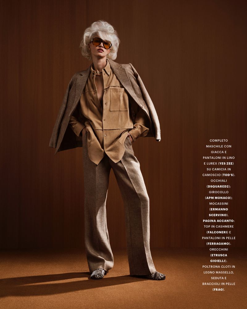 A woman poses in a tan blazer, button-up shirt, and slacks, accessorized with sunglasses and jewelry, against a solid brown backdrop in a studio shot.