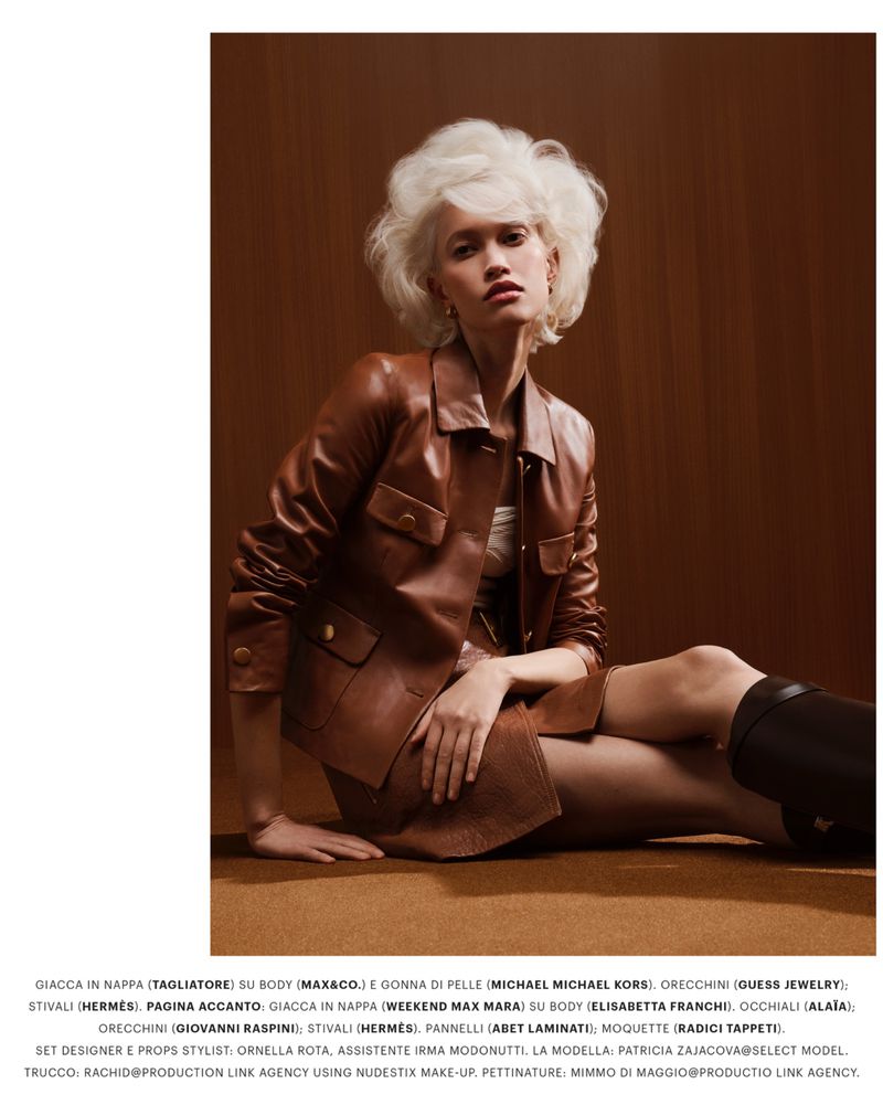 Model Patricia Zajac with voluminous white hair wearing a brown leather jacket and skirt seated against a brown wood paneling background.