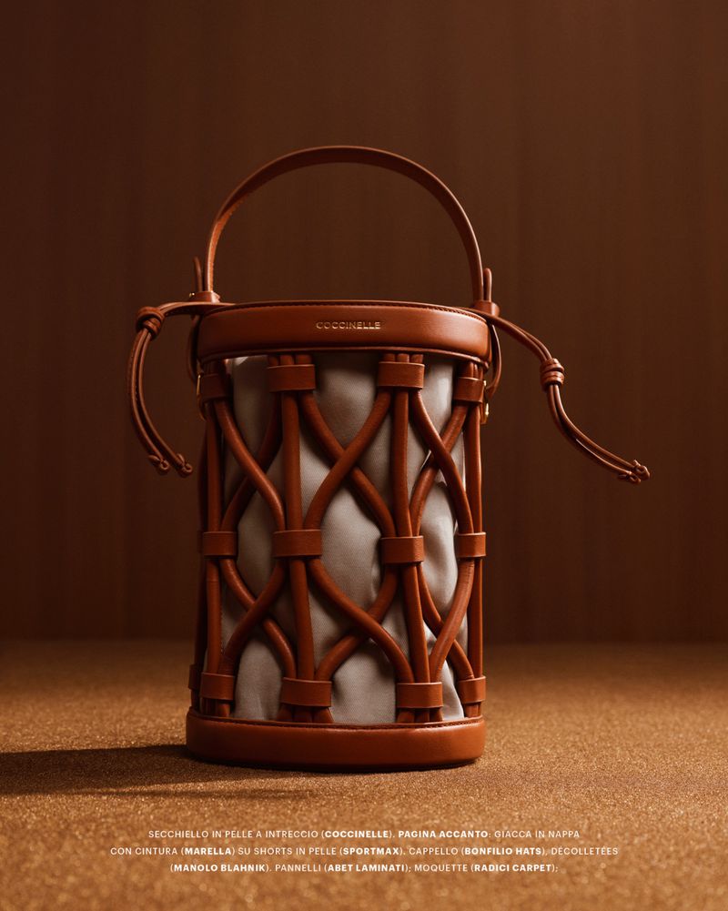 Brown leather woven bucket bag with top handle resting on a textured brown surface against a warm brown background