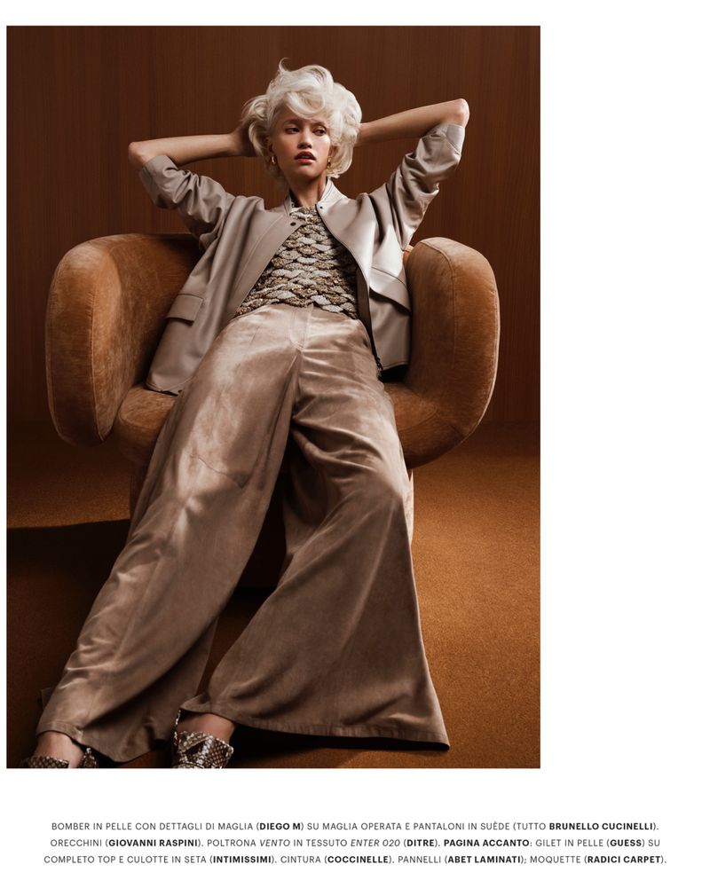 Model with platinum blonde hair seated in a brown armchair wearing a tan jacket and wide trousers for Grazia Italy.