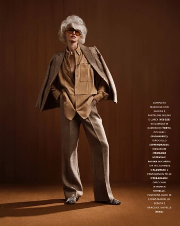Model with voluminous white hair wearing a tan button-down shirt, trousers, and a blazer draped over shoulders against a warm brown wall.
