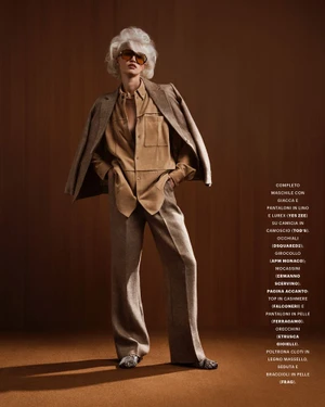 Model with voluminous white hair wearing a tan button-down shirt, trousers, and a blazer draped over shoulders against a warm brown wall.