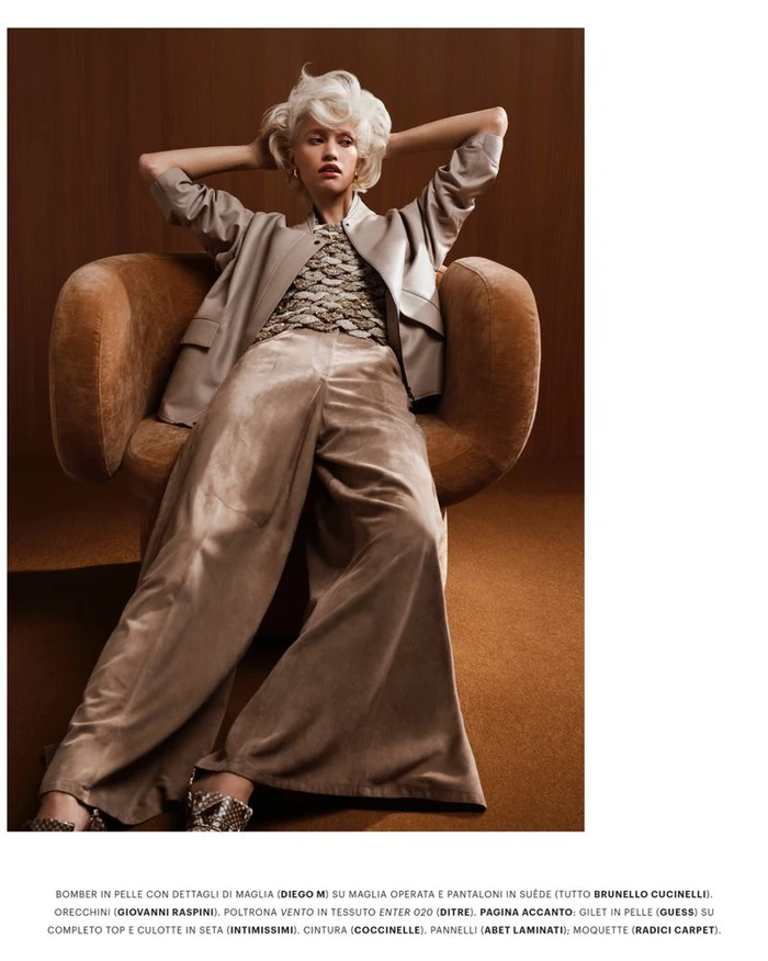 Model with platinum blonde hair seated in a brown armchair wearing a tan jacket and wide trousers for Grazia Italy.