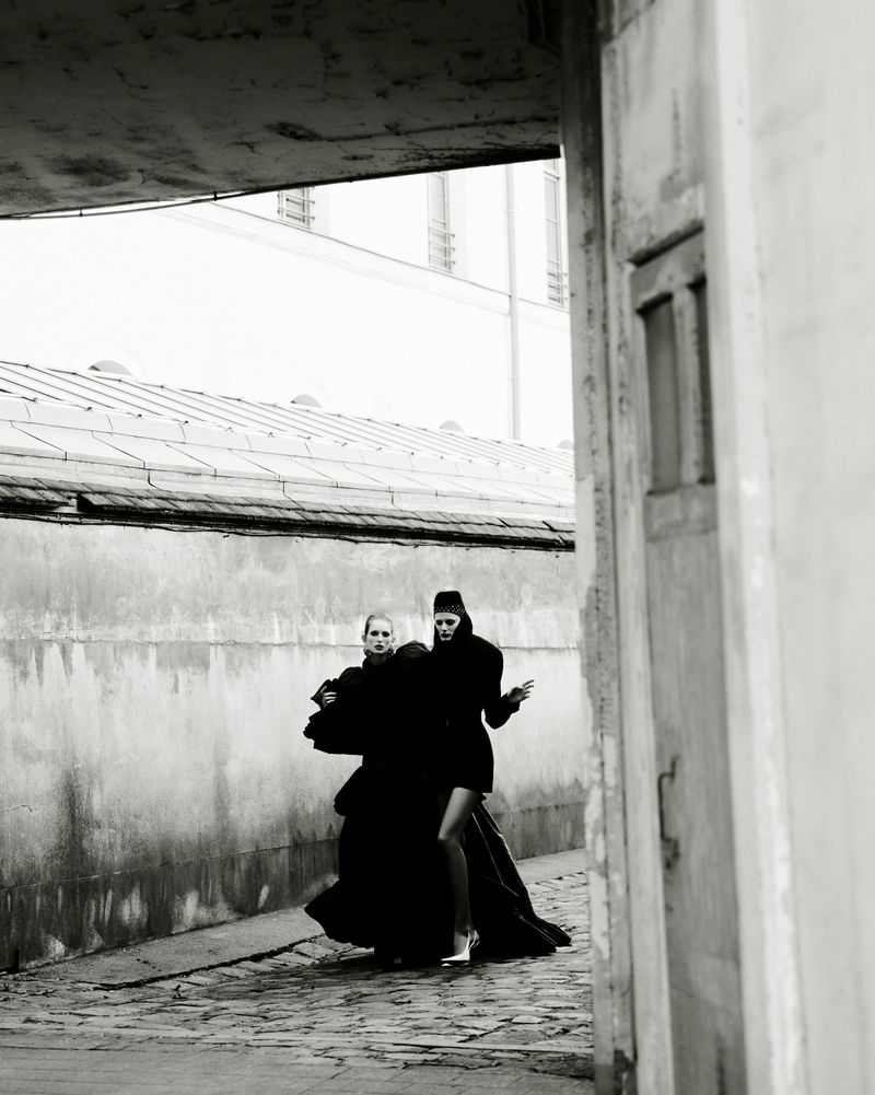 Two models in dark attire walking along an alleyway beneath an architectural overhang, photographed by Robin Galiegue.