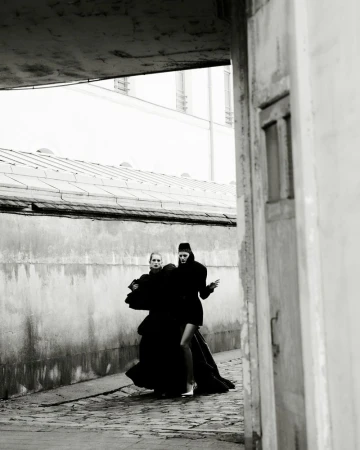 Two models in dark attire walking along an alleyway beneath an architectural overhang, photographed by Robin Galiegue.