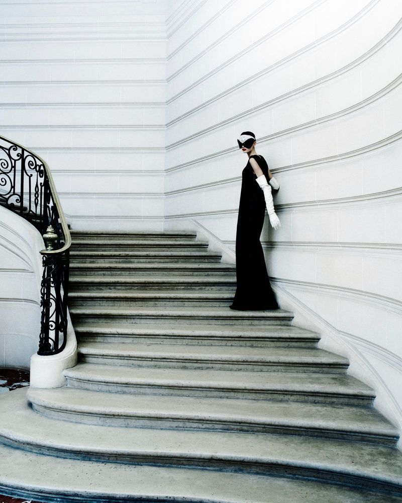 Model in a long black dress and white gloves standing on grand stone stairs against white walls