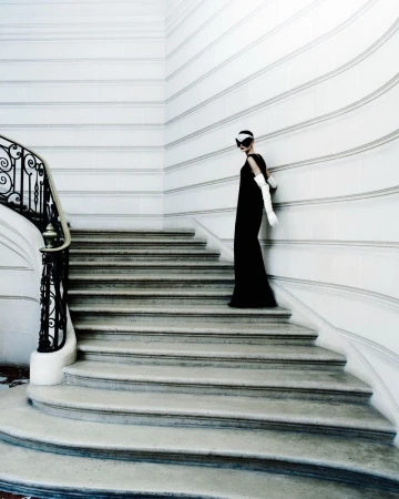 Model in a long black dress and white gloves standing on grand stone stairs against white walls