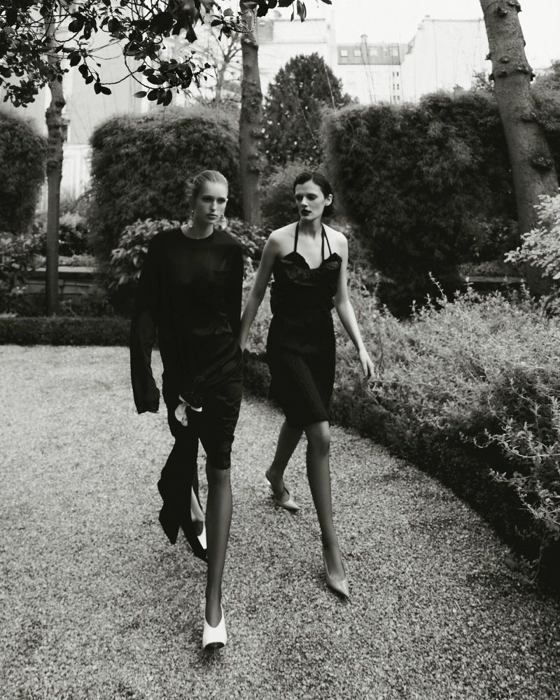 Black and white editorial image of two female models walking together along a gravel garden path flanked by hedges and trees.
