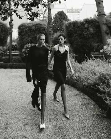 Black and white editorial image of two female models walking together along a gravel garden path flanked by hedges and trees.