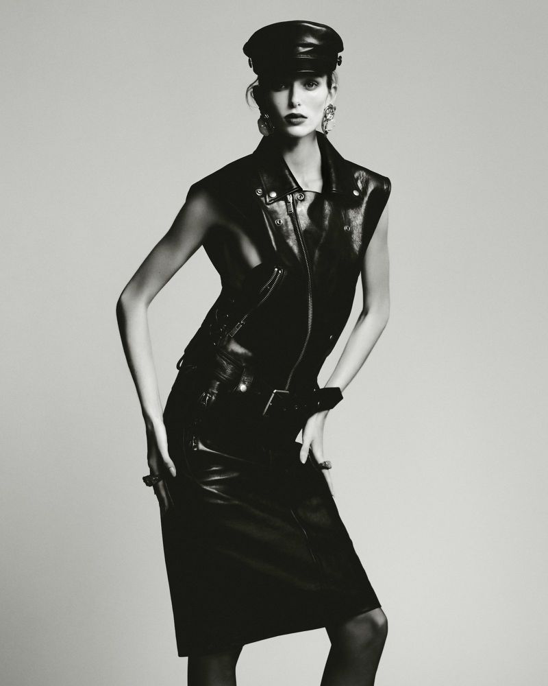 Model in black leather vest and matching skirt wearing a leather cap against a grey seamless background