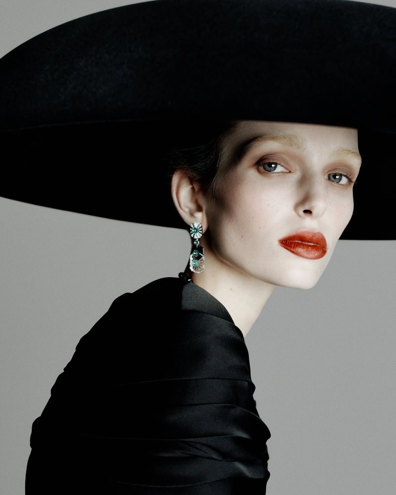 Model wearing a wide-brimmed black hat, statement earrings, and a black satin top against a grey background.