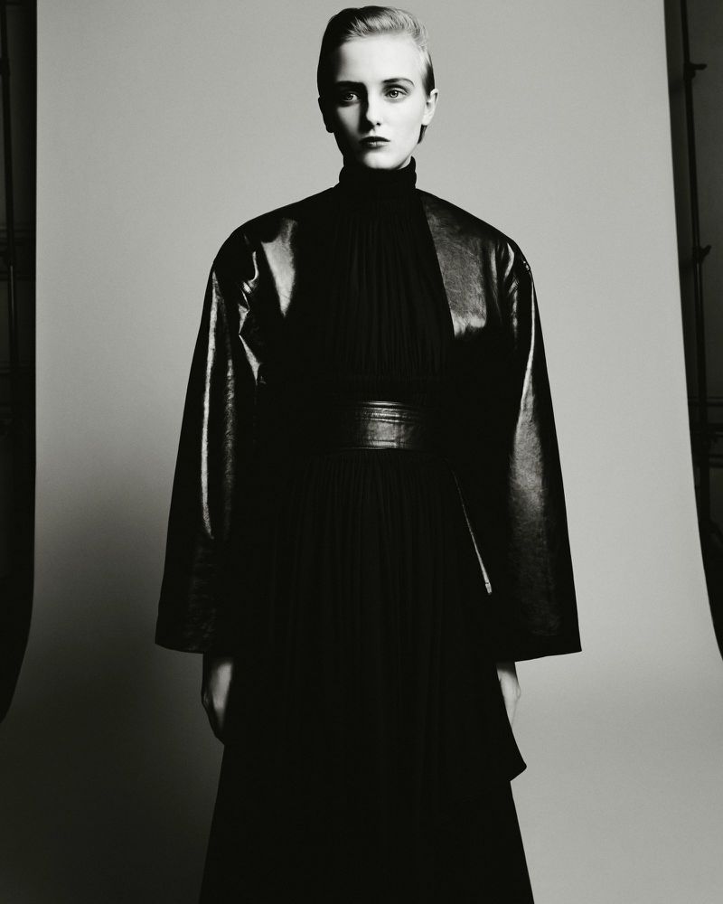 Model in black turtleneck and glossy jacket posing against a grey seamless studio background in black and white