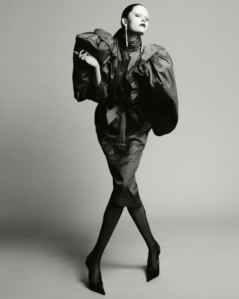 Model in dark sculptural coat wearing statement earrings against a grey studio backdrop