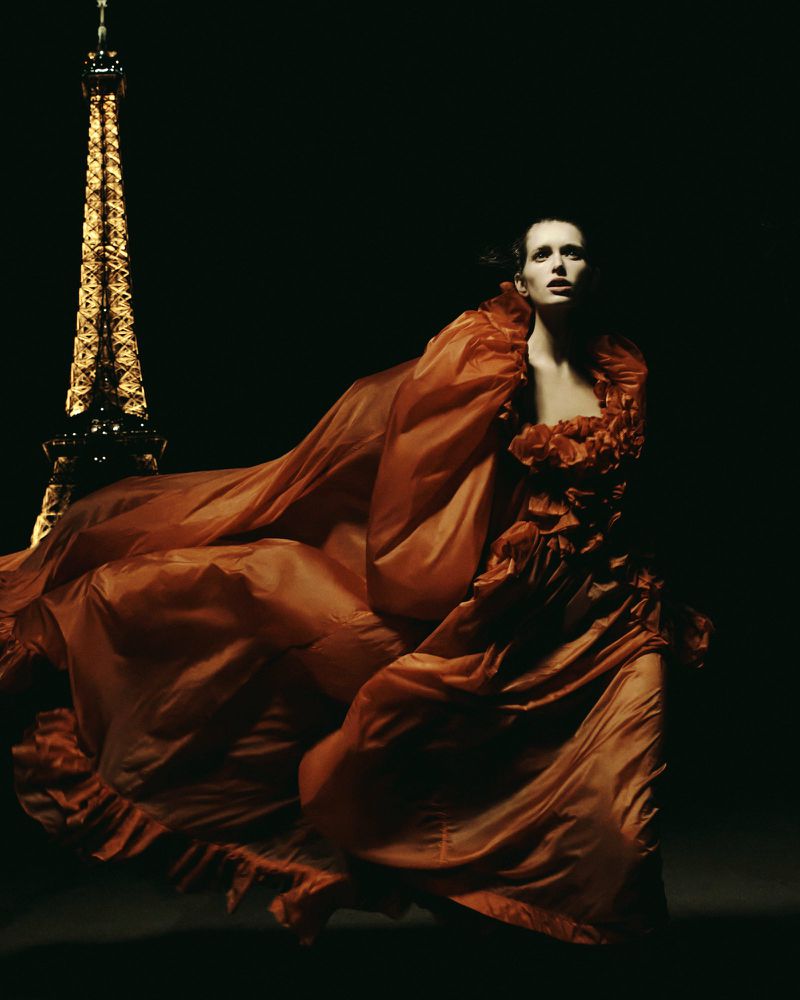 A woman in a flowing rust-colored gown poses with the Eiffel Tower in the background, creating a vintage fashion editorial style photograph.