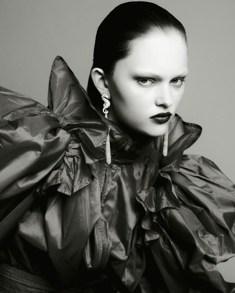 Model in ruffled dark jacket wearing statement seahorse earrings against a grey seamless backdrop