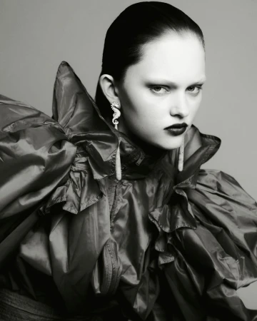 Model in ruffled dark jacket wearing statement seahorse earrings against a grey seamless backdrop