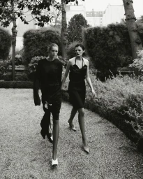Black and white editorial image of two female models walking together along a gravel garden path flanked by hedges and trees.