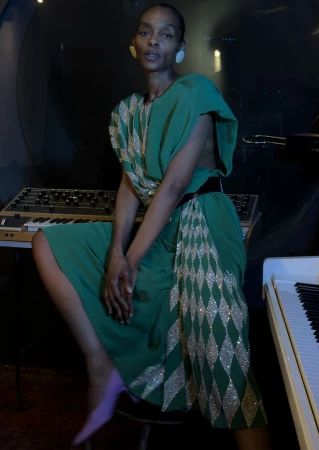 Black woman in a green dress with silver diamond patterns posing seated near a synthesizer for The Face Magazine.