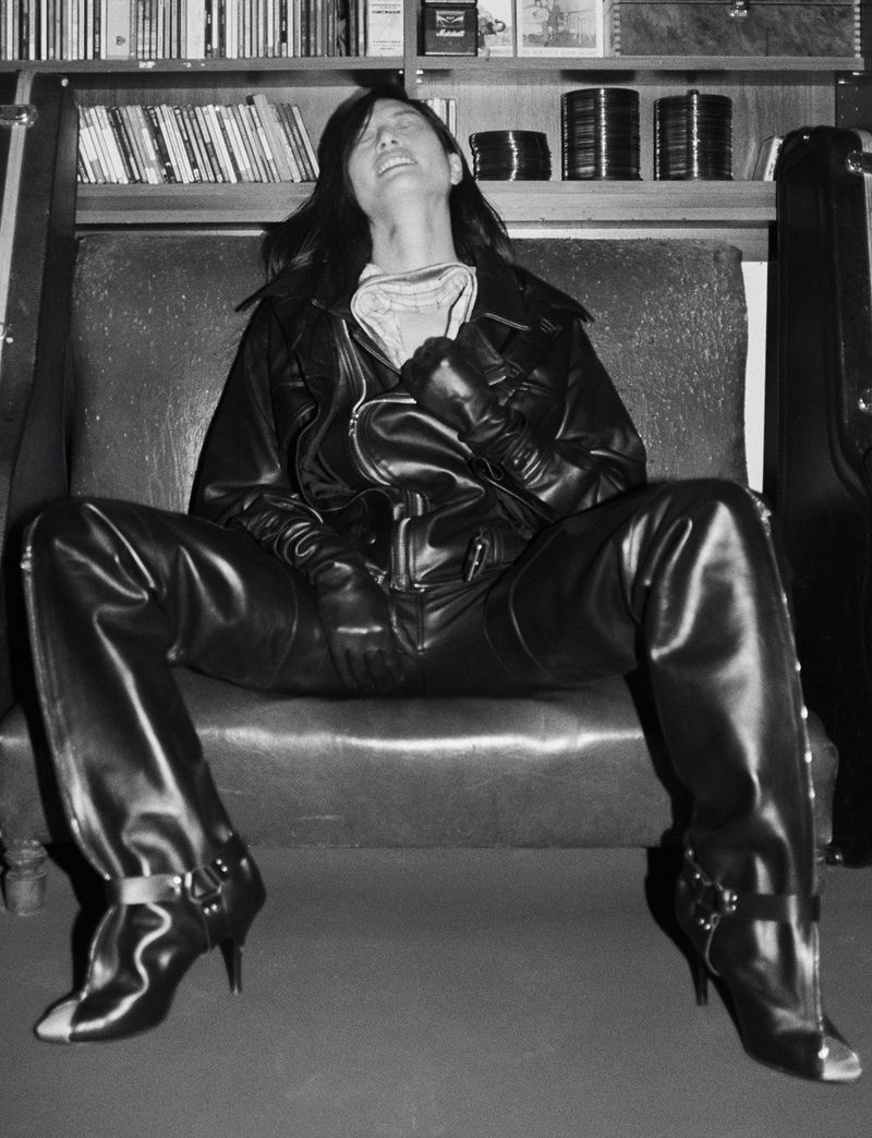 Monochrome portrait of a person in a black leather jacket and boots seated on a sofa against a bookshelf backdrop.