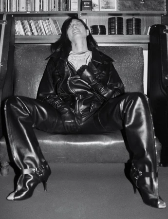 Monochrome portrait of a person in a black leather jacket and boots seated on a sofa against a bookshelf backdrop.