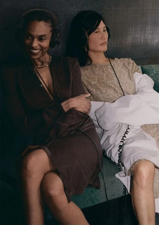 Two women wearing headphones seated next to each other on a dark textured sofa for The Face Magazine