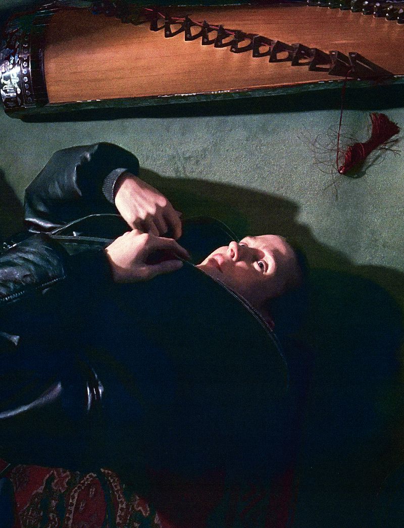 A person lying down in a dark interior space with a traditional stringed instrument positioned horizontally above them.