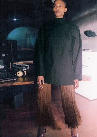 Model wearing a dark oversized top and brown fringed trousers standing in a music recording studio environment.