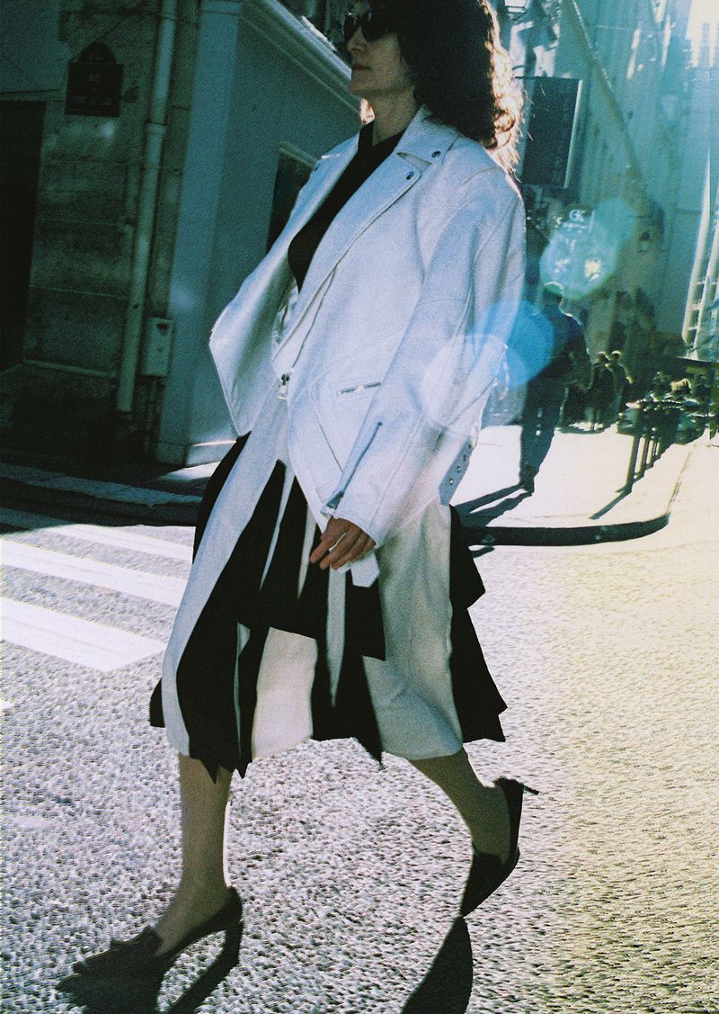 Woman walking on a sunny urban street wearing a white leather jacket and pleated skirt, captured in editorial style.