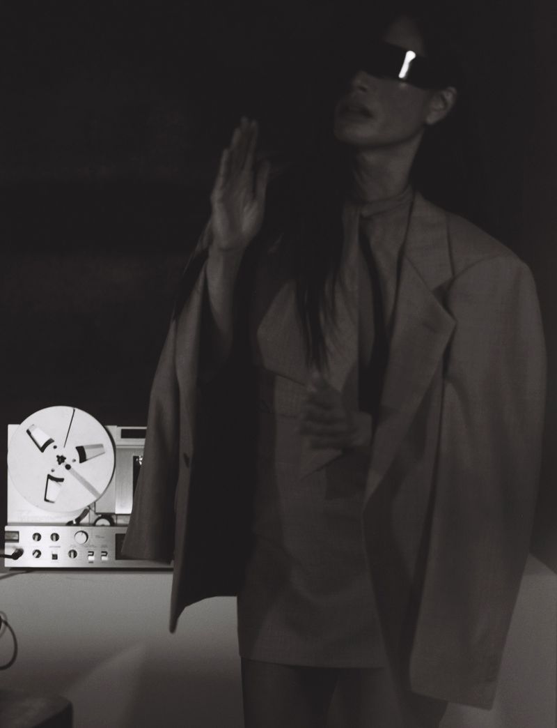 Monochrome fashion editorial portrait of a woman wearing futuristic visor sunglasses and an oversized blazer near a reel-to-reel tape recorder