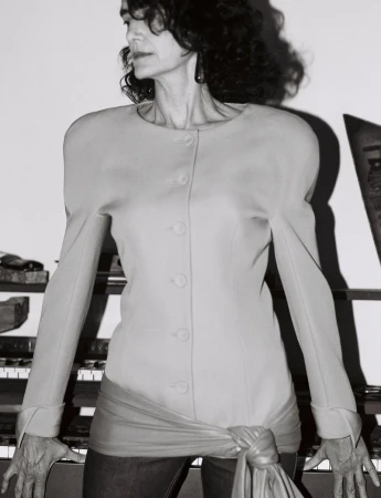 Portrait of a woman wearing a structured buttoned jacket and knotted sash standing before a piano for The Face Magazine.