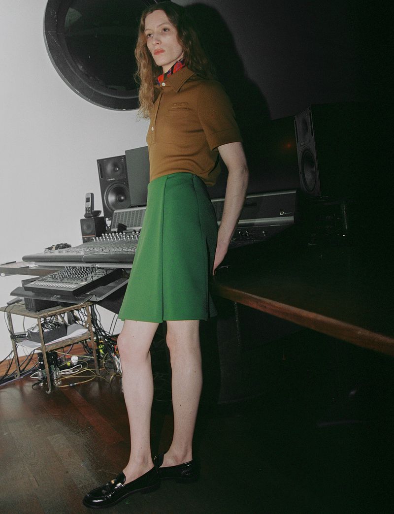 Model standing in a recording studio wearing a brown polo shirt and green skirt next to a mixing console