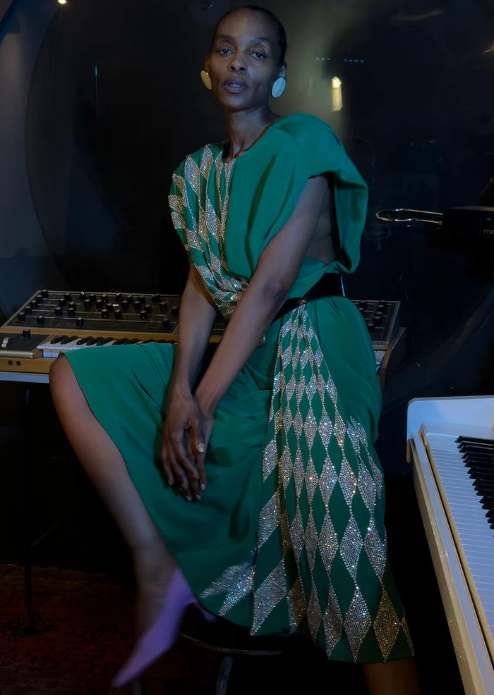 Black woman in a green dress with silver diamond patterns posing seated near a synthesizer for The Face Magazine.