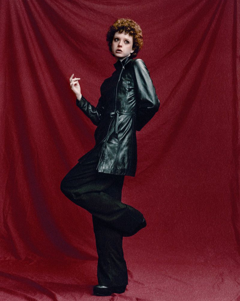 Model Mae O'Neill wearing a black leather jacket and trousers posing against a deep red fabric backdrop