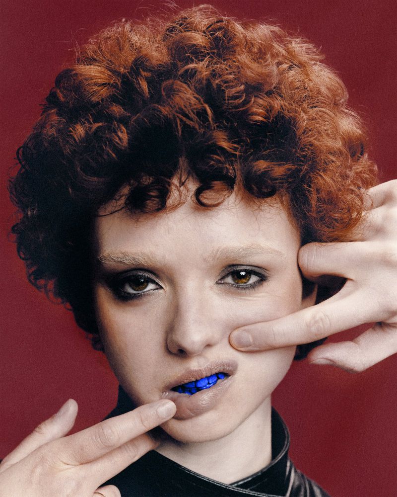 Portrait of a young woman with auburn curly hair and light skin, posed in front of a red background. Hands frame her face.