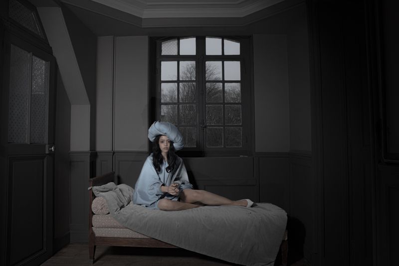 A woman in a blue dress and large matching hat seated on a daybed in a dimly lit room for Dazed MENA I'TIMAD