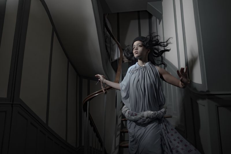 Woman in draped pale blue dress descending a dark staircase holding a chain necklace for Dazed MENA I'TIMAD project