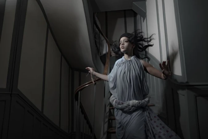 Woman in draped pale blue dress descending a dark staircase holding a chain necklace for Dazed MENA I'TIMAD project