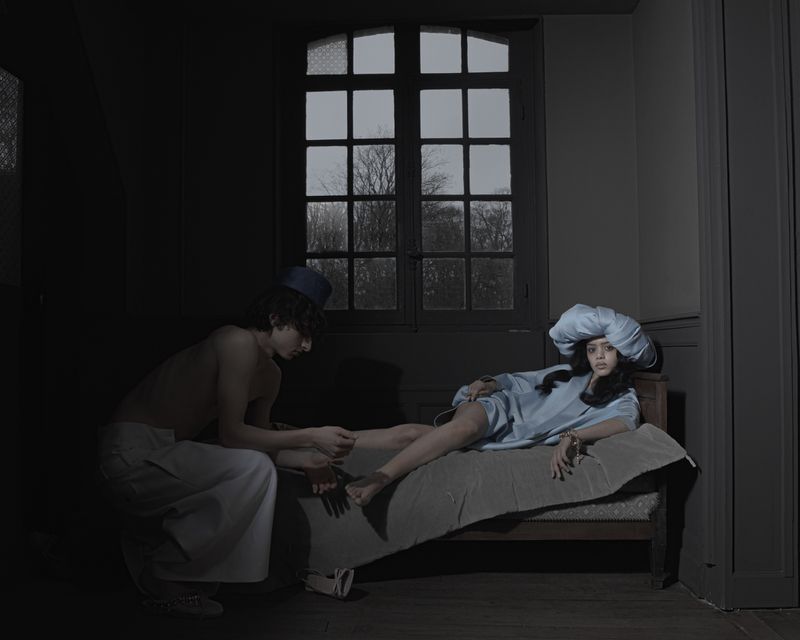 A man kneeling by a bed touching a woman's foot in a dimly lit room with a large window.