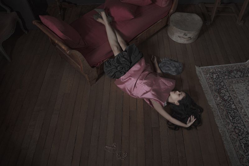 Model lying on wooden floor wearing a pink sleeveless top and dark skirt near a red daybed for I Timad project