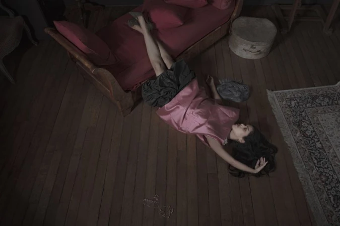 Model lying on wooden floor wearing a pink sleeveless top and dark skirt near a red daybed for I Timad project