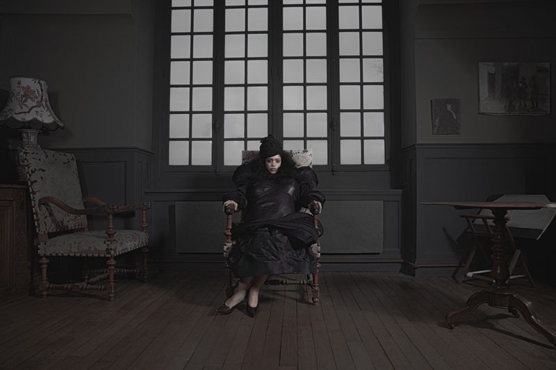 A person in dark garments seated in an armchair inside a dimly lit room with a large grid window and dark walls.
