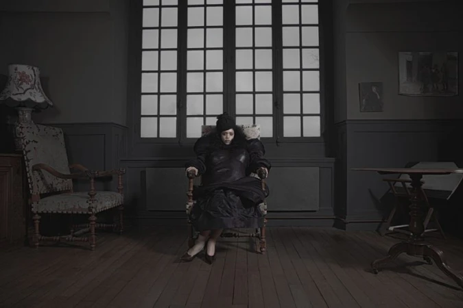 A person in dark garments seated in an armchair inside a dimly lit room with a large grid window and dark walls.