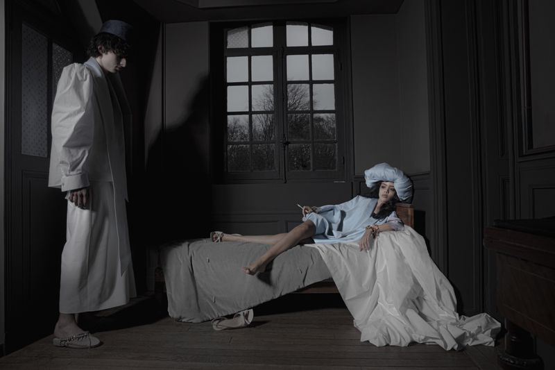 Man in white tunic standing near a woman lying on a draped bed in a dark room for Dazed MENA I'TIMAD