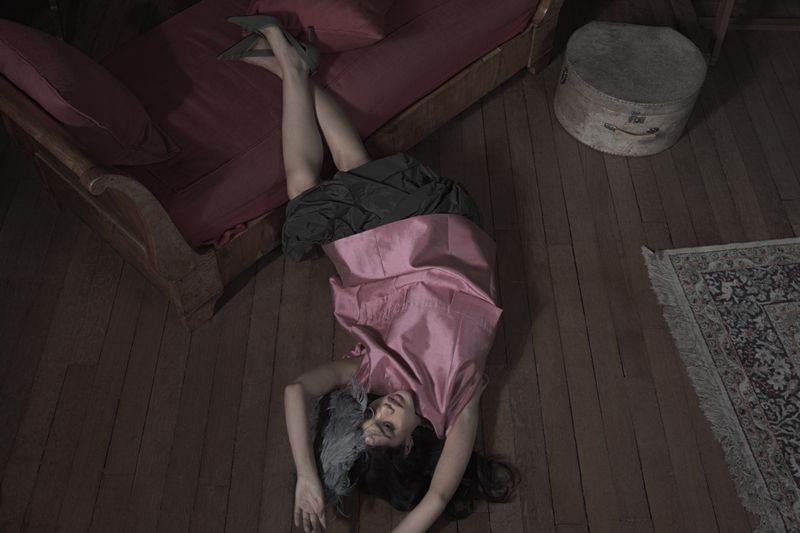 Model wearing a pink top and dark skirt lying on a wooden floor beside a red couch for Dazed MENA project I’TIMAD.