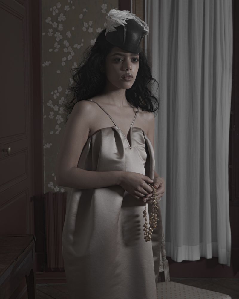 Woman wearing a satin dress and feathered headpiece holding a beaded necklace indoors for Dazed MENA I'TIMAD