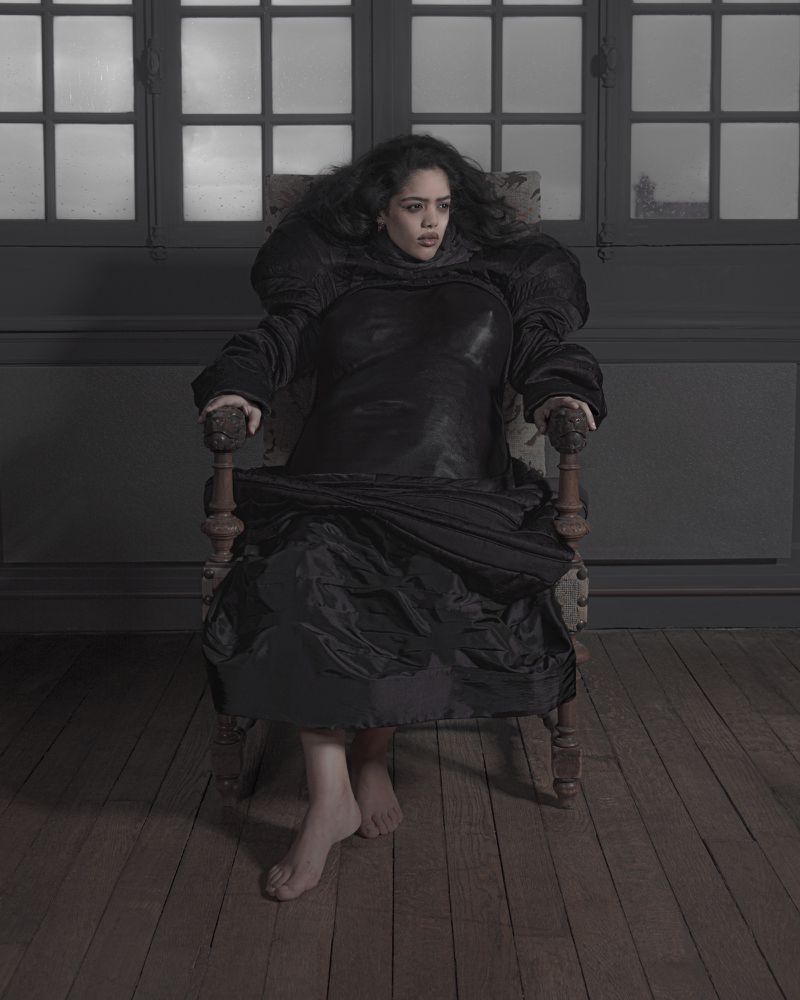 Woman seated barefoot in an antique armchair wearing a dark voluminous garment in a room with gridded window panes
