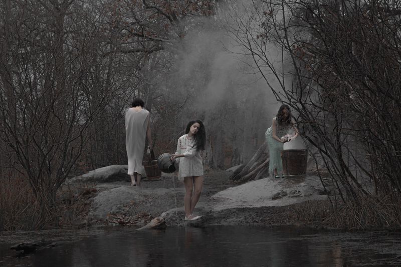 Three women near a pond in an atmospheric outdoor setting, wearing light-colored dresses and carrying buckets, creating a sense of eerie.