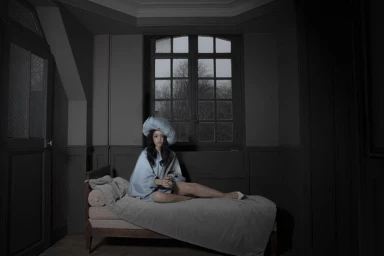 A woman in a blue dress and large matching hat seated on a daybed in a dimly lit room for Dazed MENA I'TIMAD