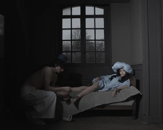 A man kneeling by a bed touching a woman's foot in a dimly lit room with a large window.