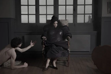 Woman seated in an ornate armchair wearing dark structured fashion, with a man kneeling beside her in an interior room.